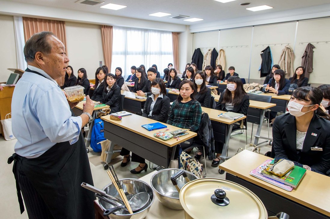 An instructor teaches Japanese cuisine to students seated at desks in a classroom at Japan Hotel School.