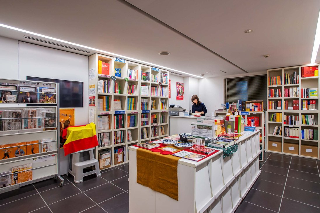 Library at Tokyo's Instituto Cervantes with bookshelves, Spanish flag, and staff member at desk.