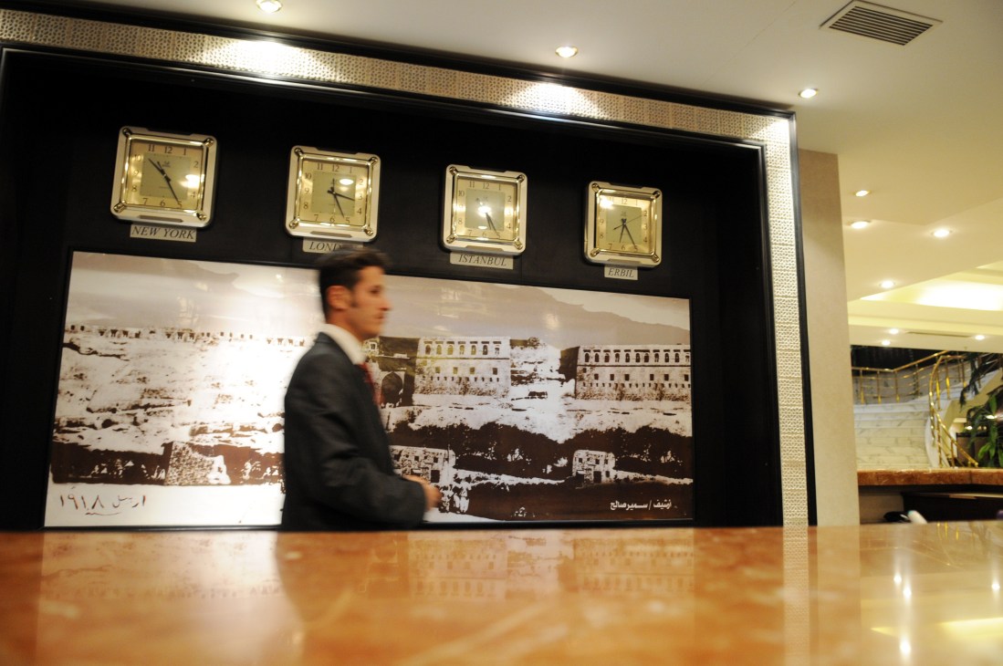 Man in dark suit stands at check-in desk with world clocks and historical photograph on wall behind.