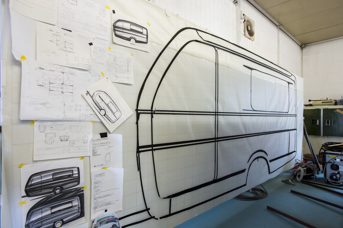 A white trailer prototype with black frame lines in a workshop, surrounded by technical drawings pinned to the wall.