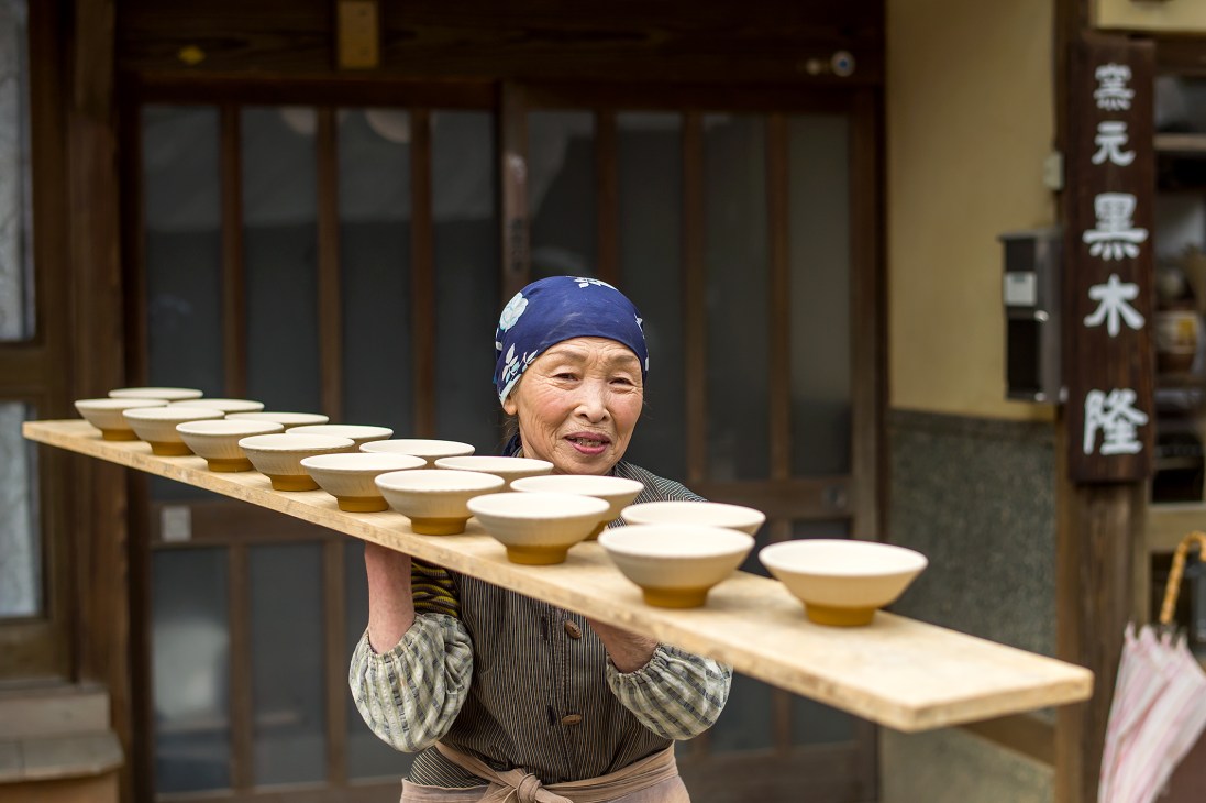 A woman in traditional dress carries a wooden board of unfinished Ontayaki pottery bowls.