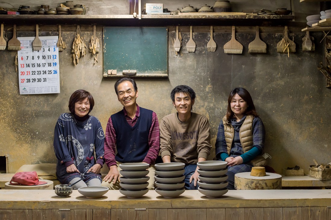 The Sakamoto family smiles at their pottery workshop counter displaying stacked ceramic bowls.