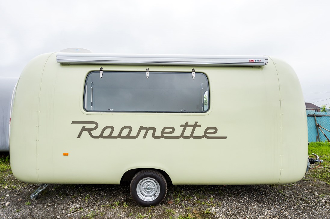 Cream-colored Roomette trailer with rounded design and large window, parked on gravel.