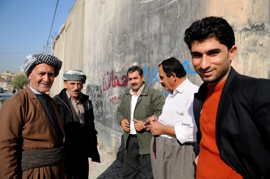 Six men standing on a street in Arbil, with traditional Kurdish dress and modern clothing, against a weathered wall with graffiti.