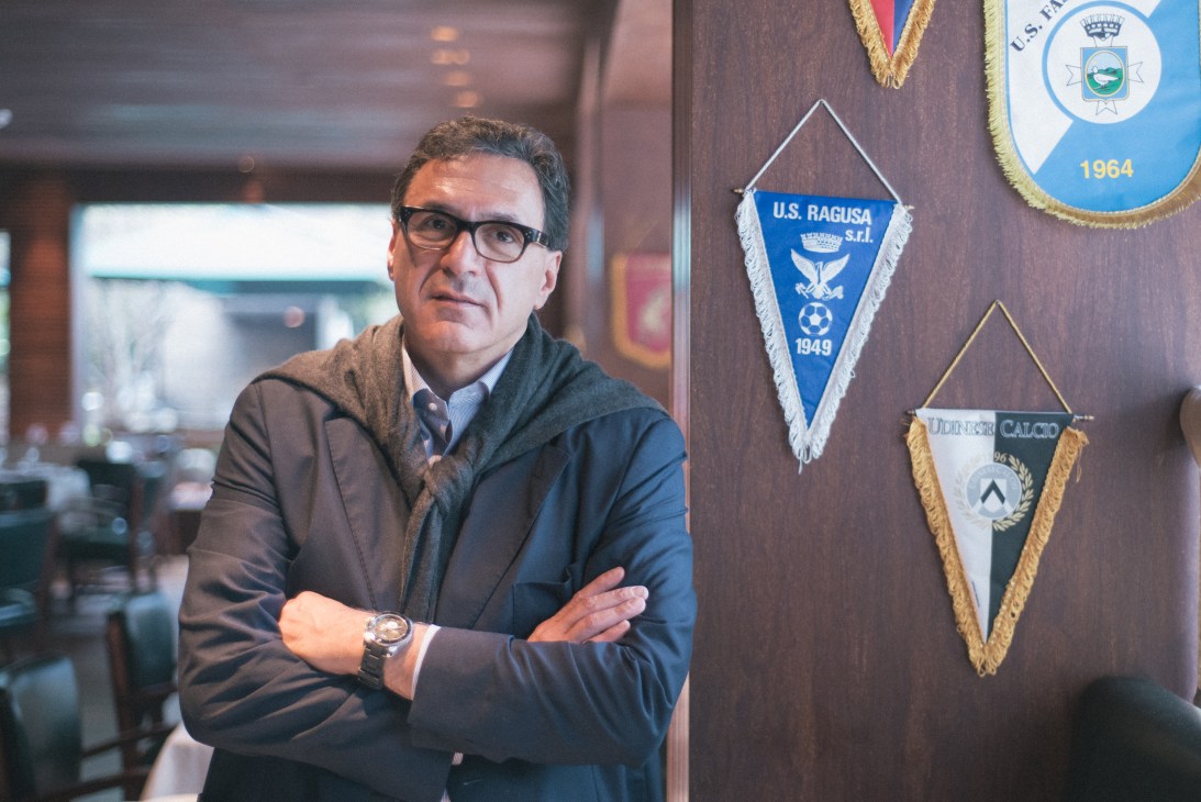 Rogério Fasano stands with arms crossed in his Trattoria restaurant beside sporting pennants on the wall.