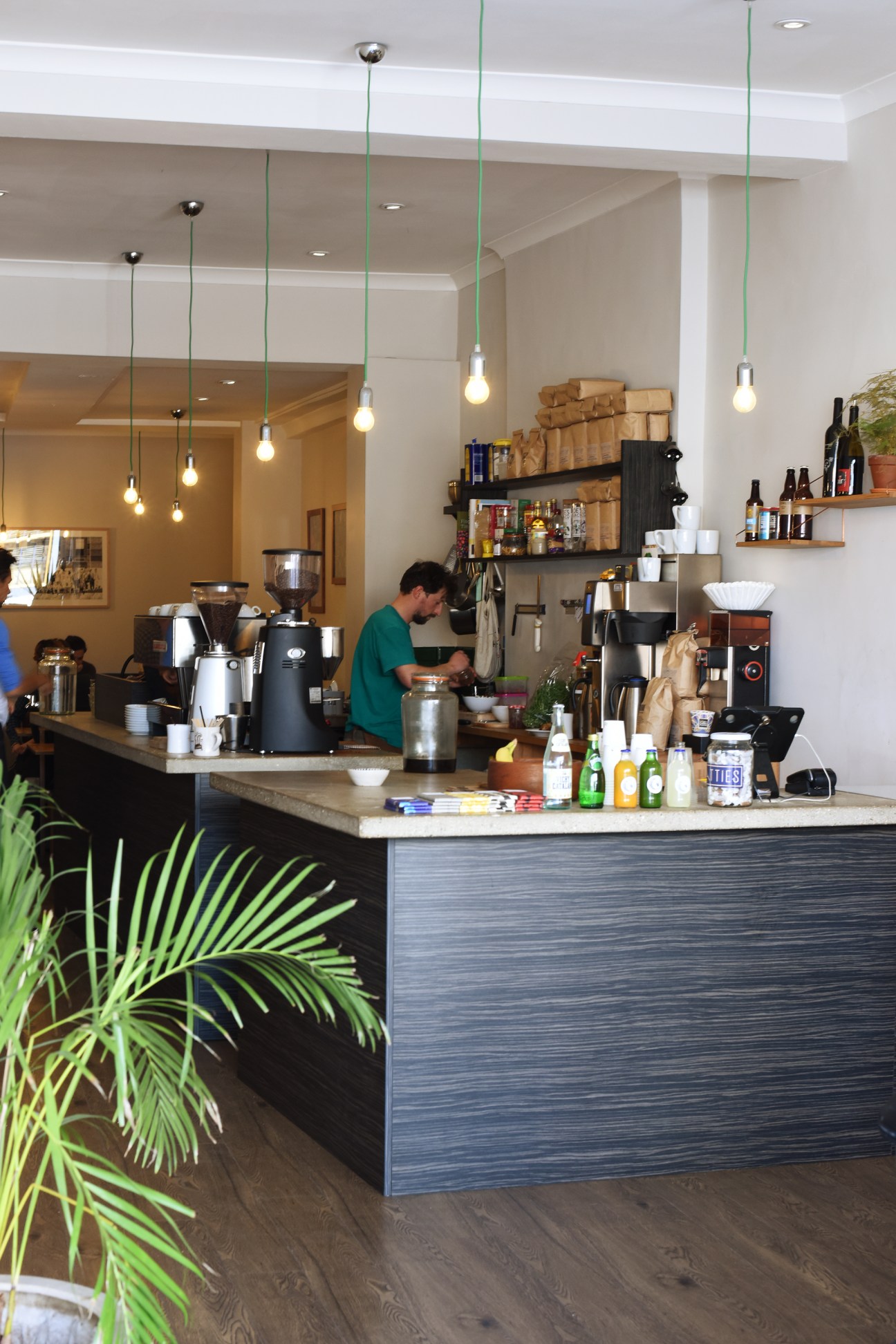 Daily Goods coffee shop interior in Camberwell with barista at counter, green pendant lights, and shelving.