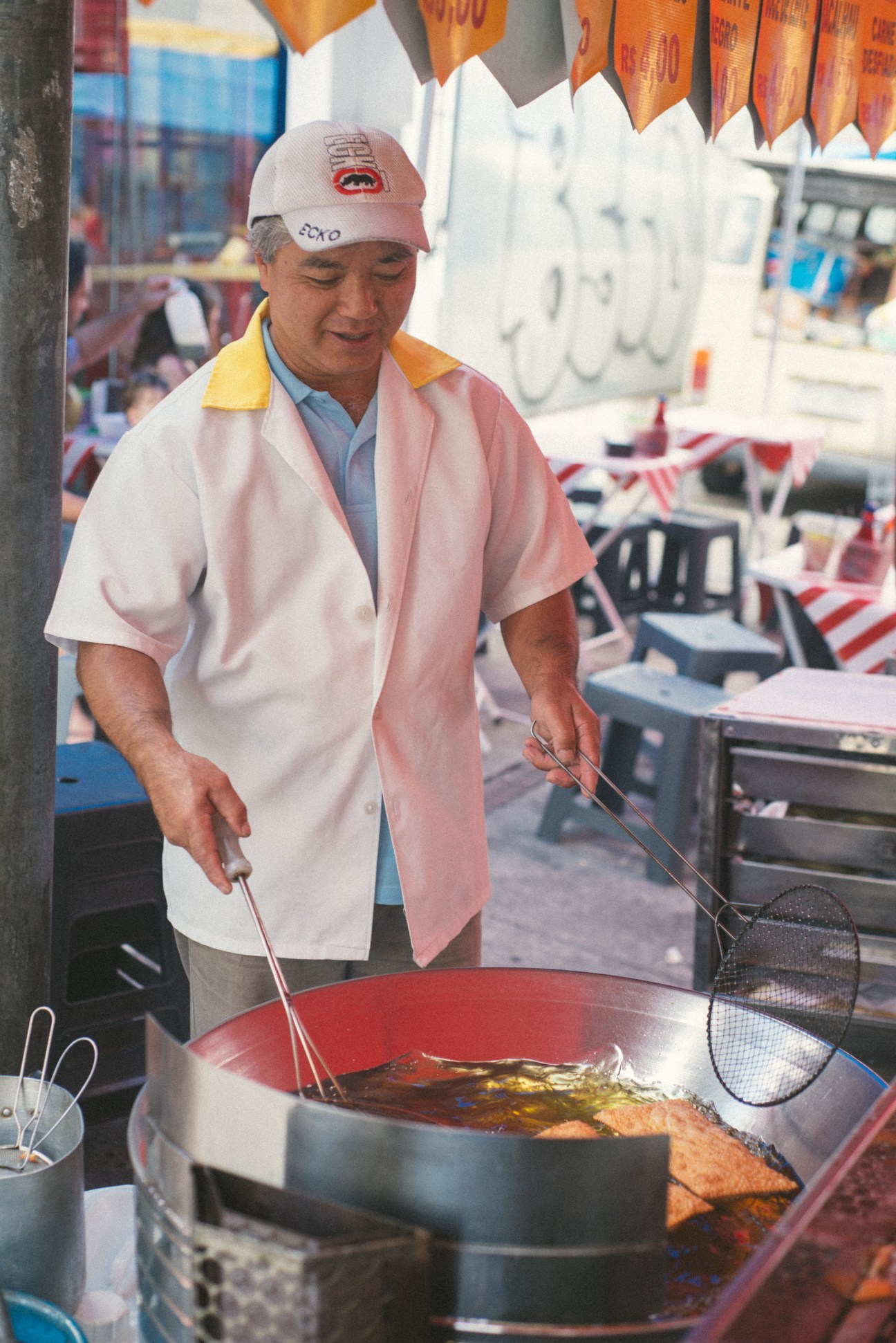 Street vendor in pink apron preparing pasteis in a red pan at a food stall.