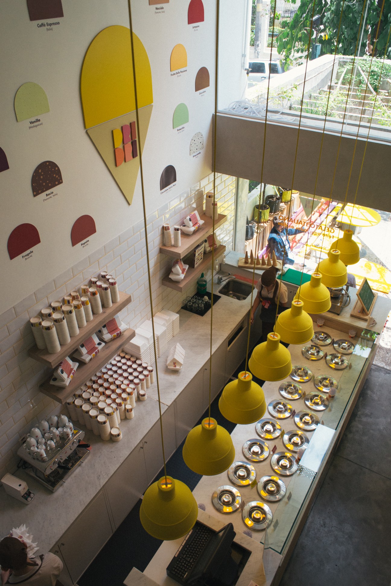 Interior of Dri Dri ice cream shop with yellow pendant lamps, flavor samples, and colorful branding on walls.