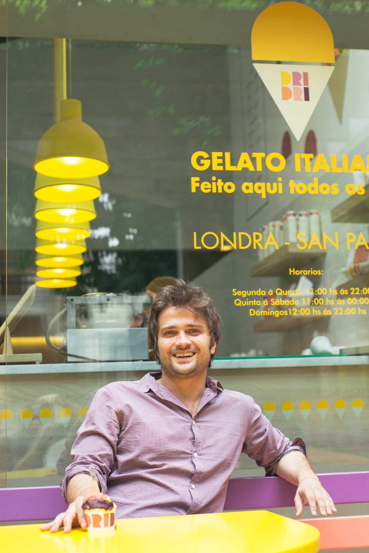 Nico de Virieu smiles outside his Dri Dri gelato shop in São Paulo, with yellow pendant lights and colorful signage visible behind him.