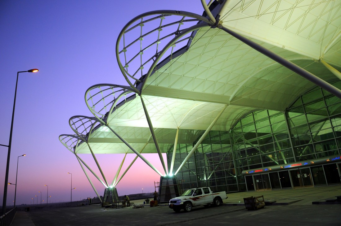 Modern illuminated airport terminal at dusk with sculptural canopy architecture and vehicle parked outside.