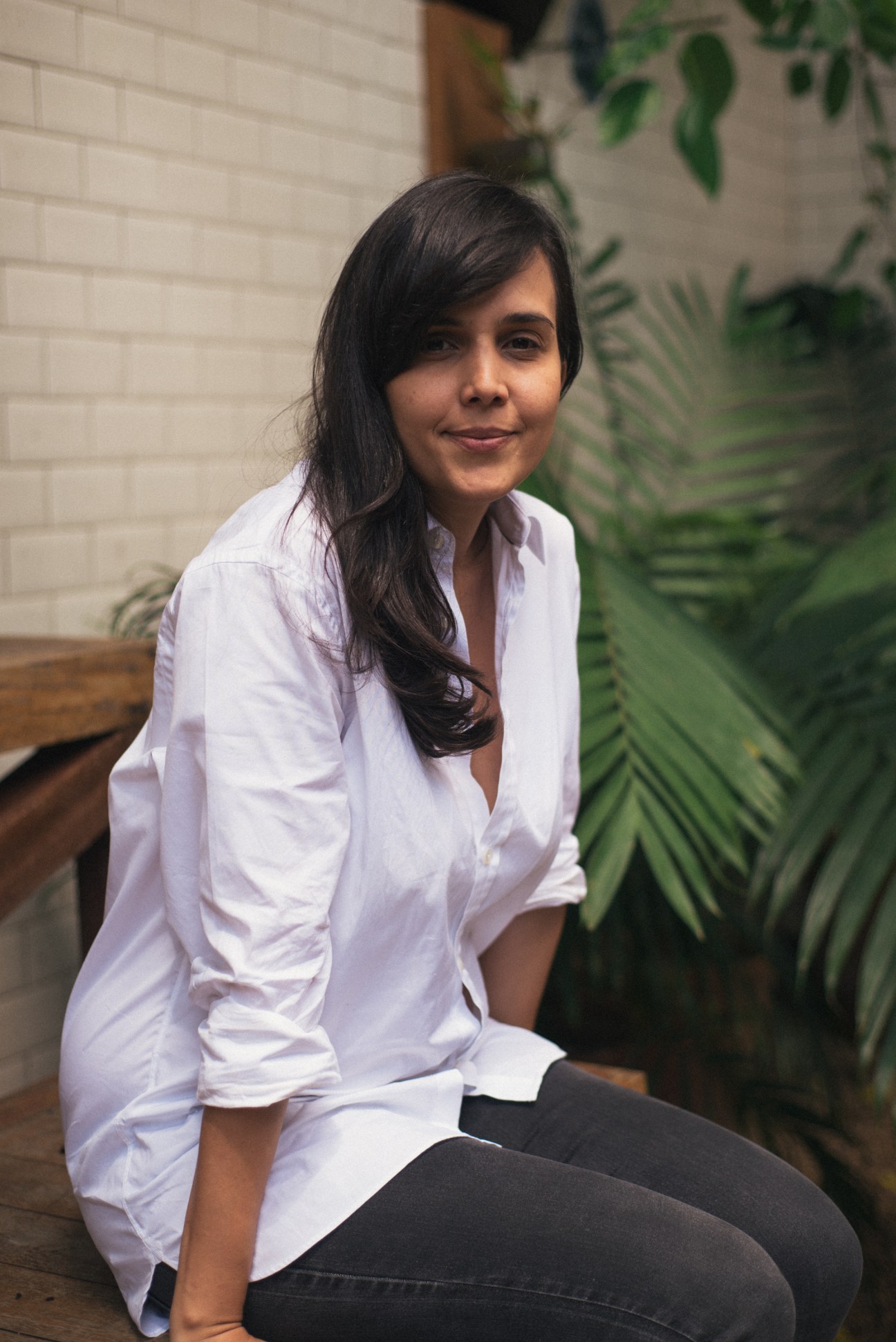 Karina Mota, owner of Chez Oscar, sits against a white brick wall with green plants.