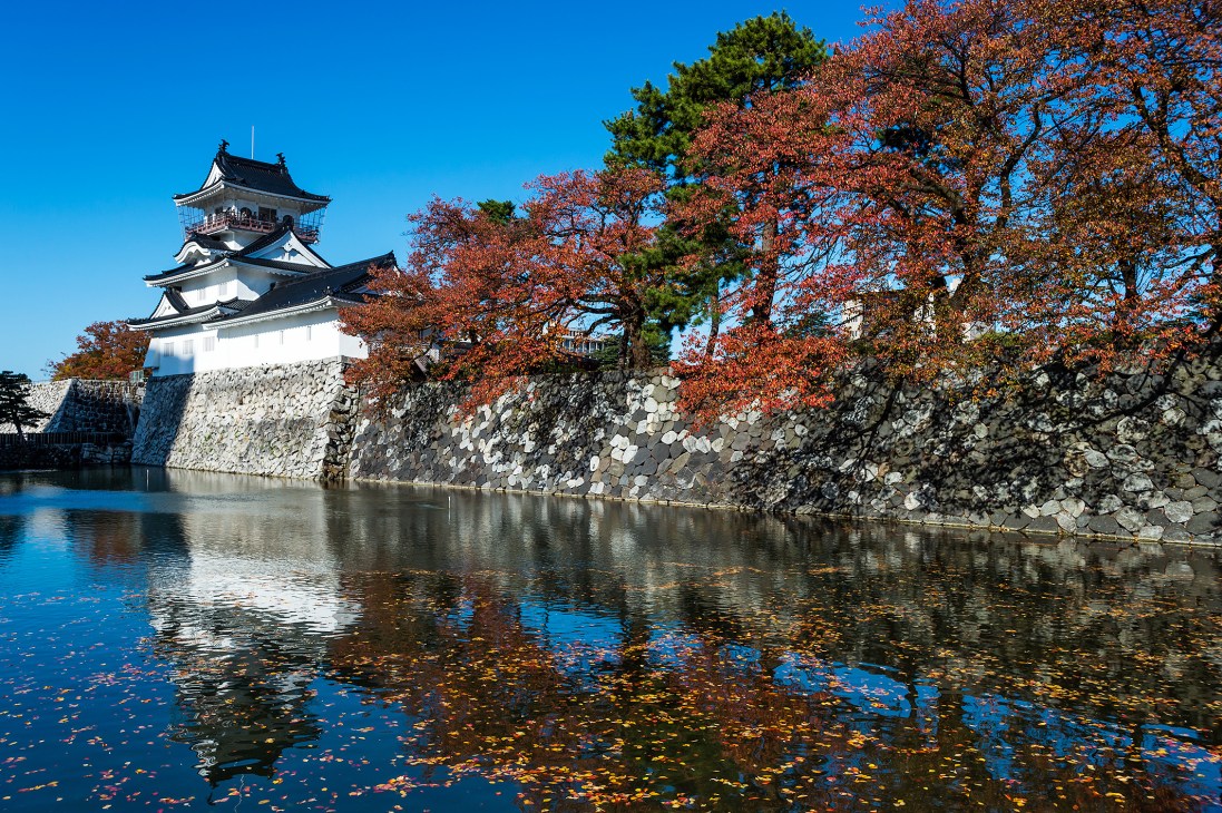 Toyama Castle with white walls and traditional tiered roofs reflected in water surrounded by autumn foliage.
