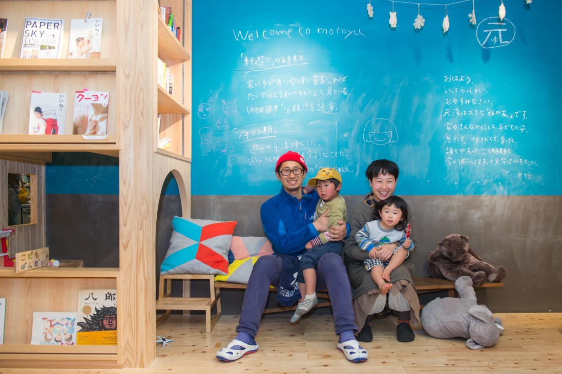 A man and woman with two young children sit on a bench in a modern interior space with a bright blue chalkboard wall covered in Japanese writing.