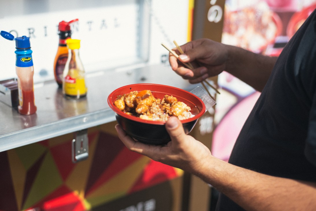 Person holding a red bowl of teriyaki chicken and rice with chopsticks at a food truck counter.