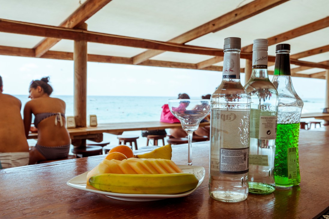 Beach bar counter with spirits bottles, cocktail glass, and fruit plate overlooking Mediterranean sea.