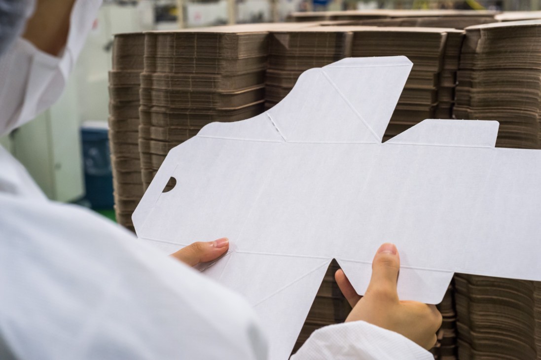 Person in white clothing examining a white cardboard box with die-cut handles against wooden backdrop.