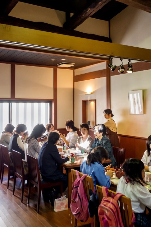 A group of seniors and other diners enjoy lunch together at a traditional Japanese restaurant with wooden beams and warm lighting.