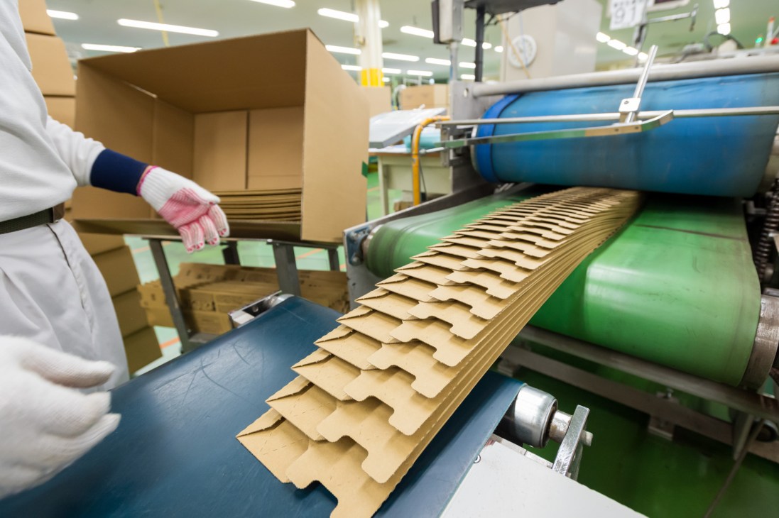 Worker in white coat and pink gloves inspecting thin cardboard packaging sheets on green industrial machinery at Crown Package factory.