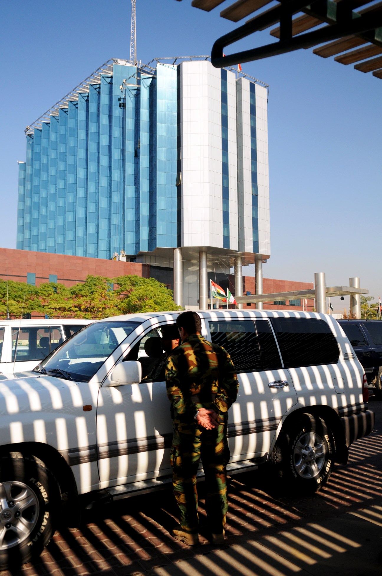 Security personnel standing beside a white SUV outside a modern glass and steel building in Arbil.