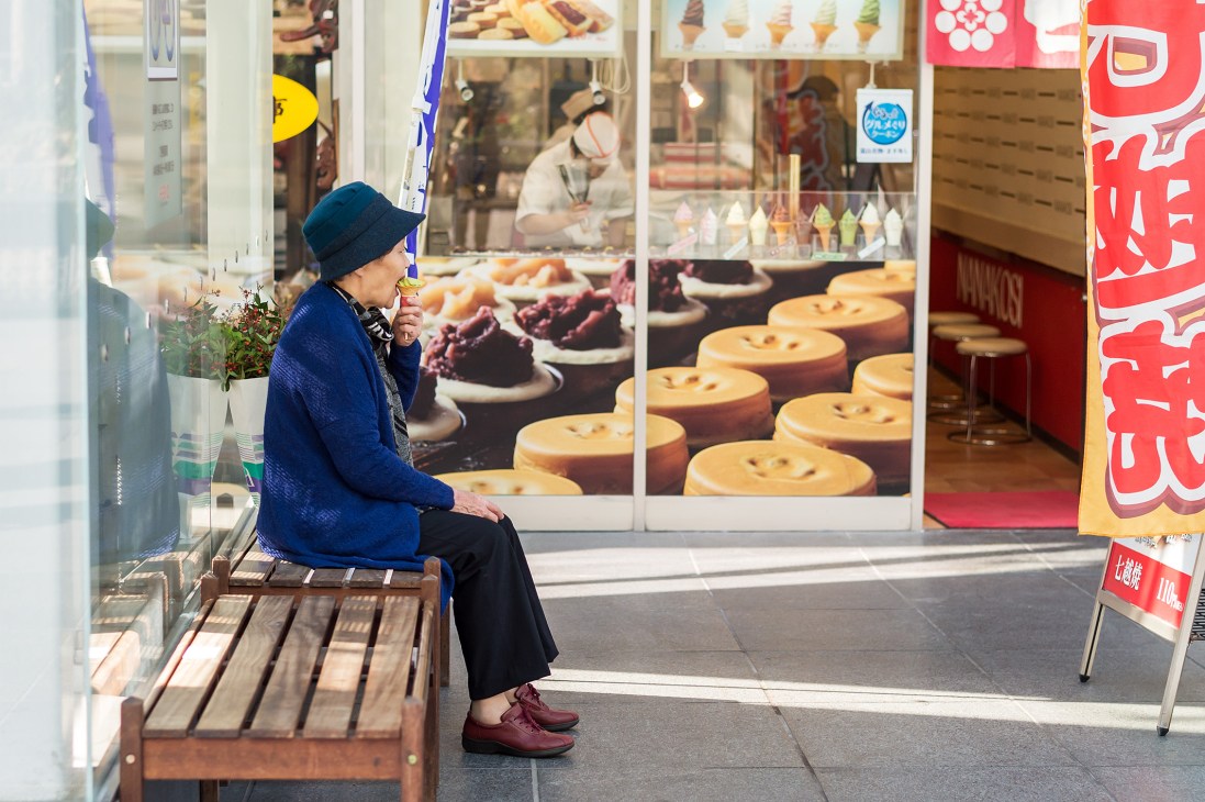 An elderly woman in a blue jacket and dark hat sits on a wooden bench outside a Japanese storefront with food display windows.