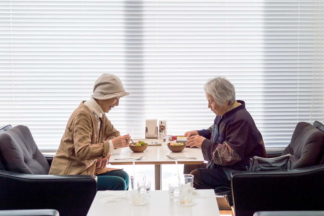 Two elderly people dining together at a table with horizontal blinds visible behind them at D & Department in Toyama.