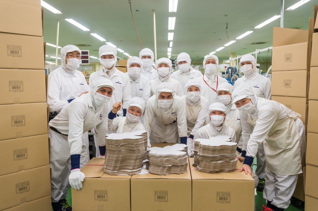 Crown Package workers in white protective uniforms and masks pose together in a packaging facility surrounded by cardboard boxes.