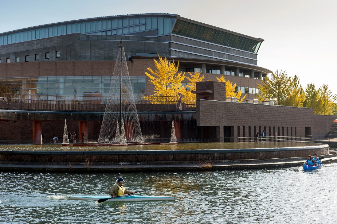 Modern waterfront building with brick and metal facade beside canal with kayakers and decorative cone structures.