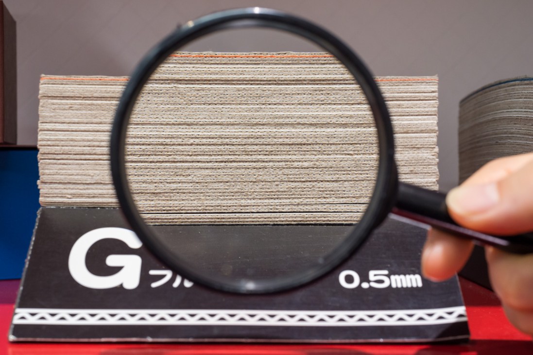 Magnifying glass showing thin corrugated cardboard material labeled 0.5mm, demonstrating Crown Package's TKP innovation.