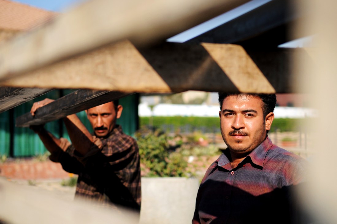 Two men at a construction site in Arbil, with one carrying wooden beams and another in the foreground.