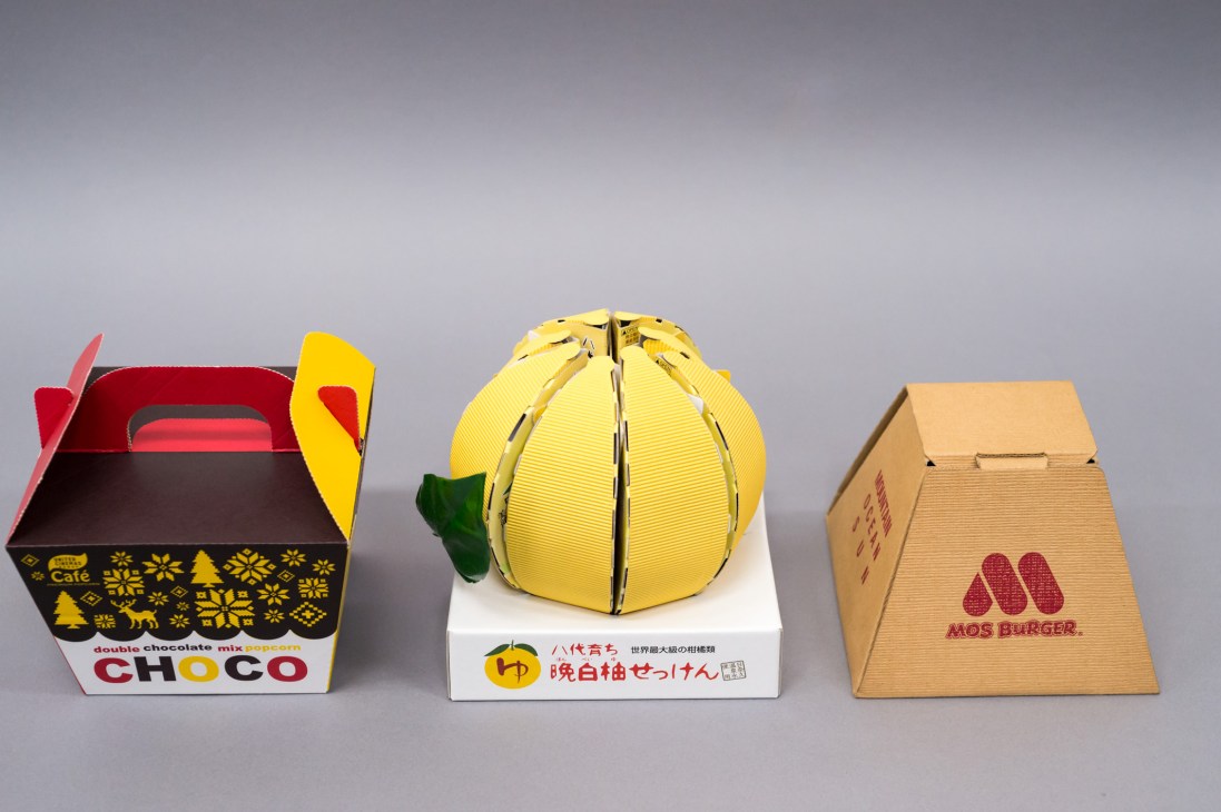 Three custom-designed packaging examples from Crown Package including a red and black box with yellow flaps, a yellow spherical citrus-shaped package, and a brown cardboard box.