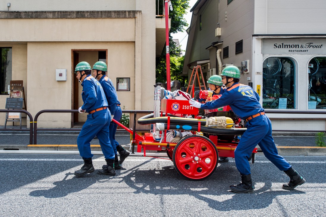 Three volunteer firefighters in blue uniforms and green helmets demonstrate equipment handling during a training exercise on a Tokyo street.