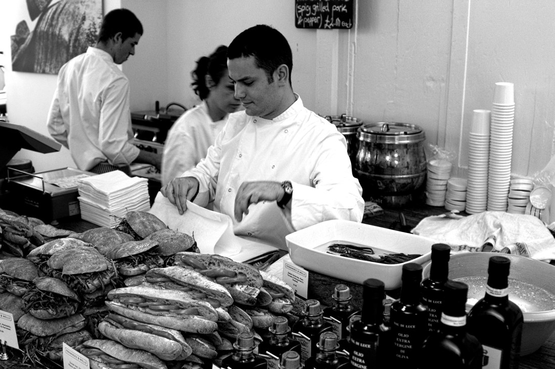 Chef in white coat preparing food at a deli counter with cured meats, breads, and olive oil bottles displayed.