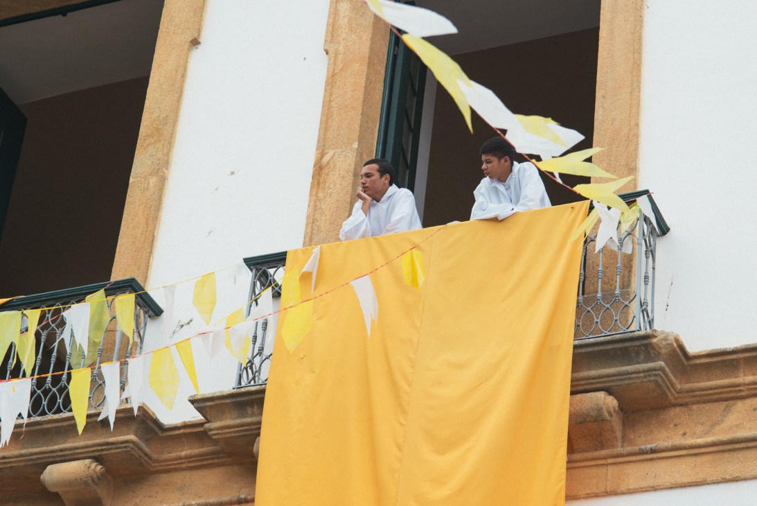 Two altar boys in white shirts stand on a colonial balcony decorated with yellow and white bunting in Paraty.