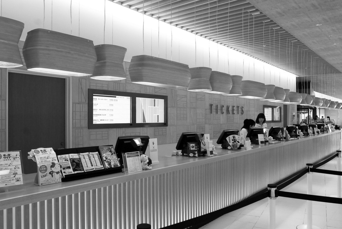 Black and white photograph of United Cinemas ticket counter with curved pendant lights and staff at registers.
