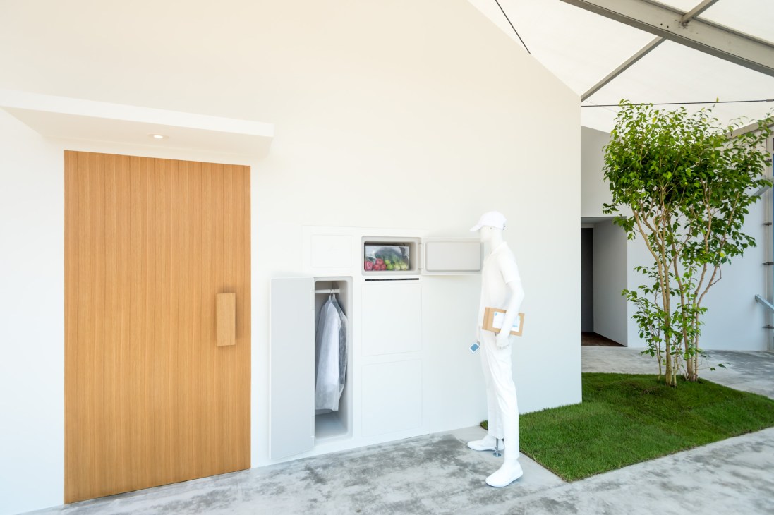 Modern white structure with external refrigerator access, wooden door, and integrated delivery system in an open courtyard with green lawn.