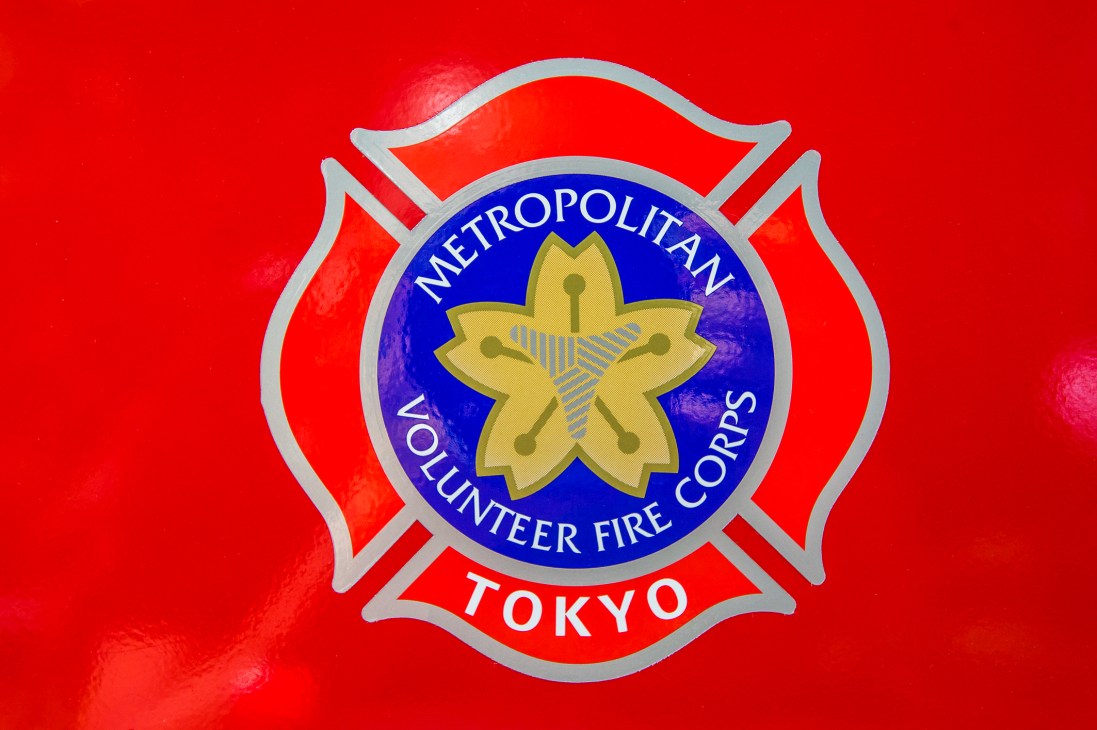 Metropolitan Volunteer Fire Corps Tokyo logo on red background with blue circular center and gold flower emblem.