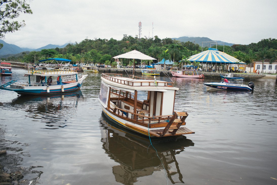 Colorful fishing boats moored in Paraty's harbor with striped carnival tents and forested mountains in the background.