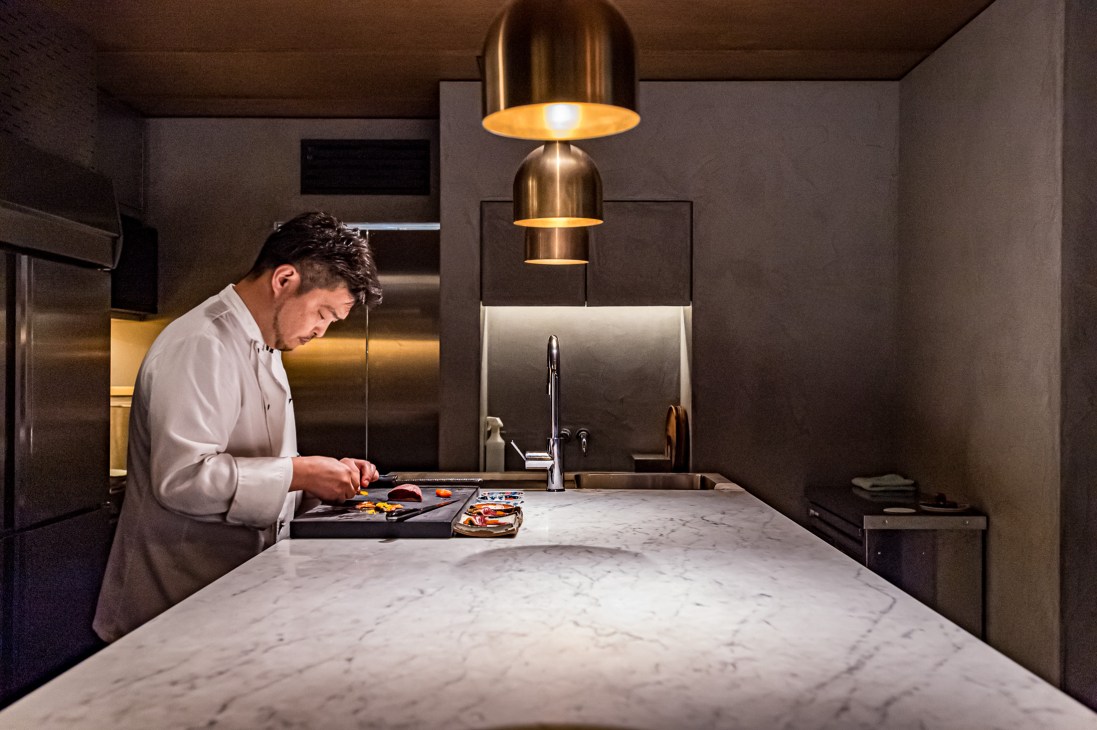 Chef preparing food at marble counter in modern kitchen with brass pendant lights overhead.