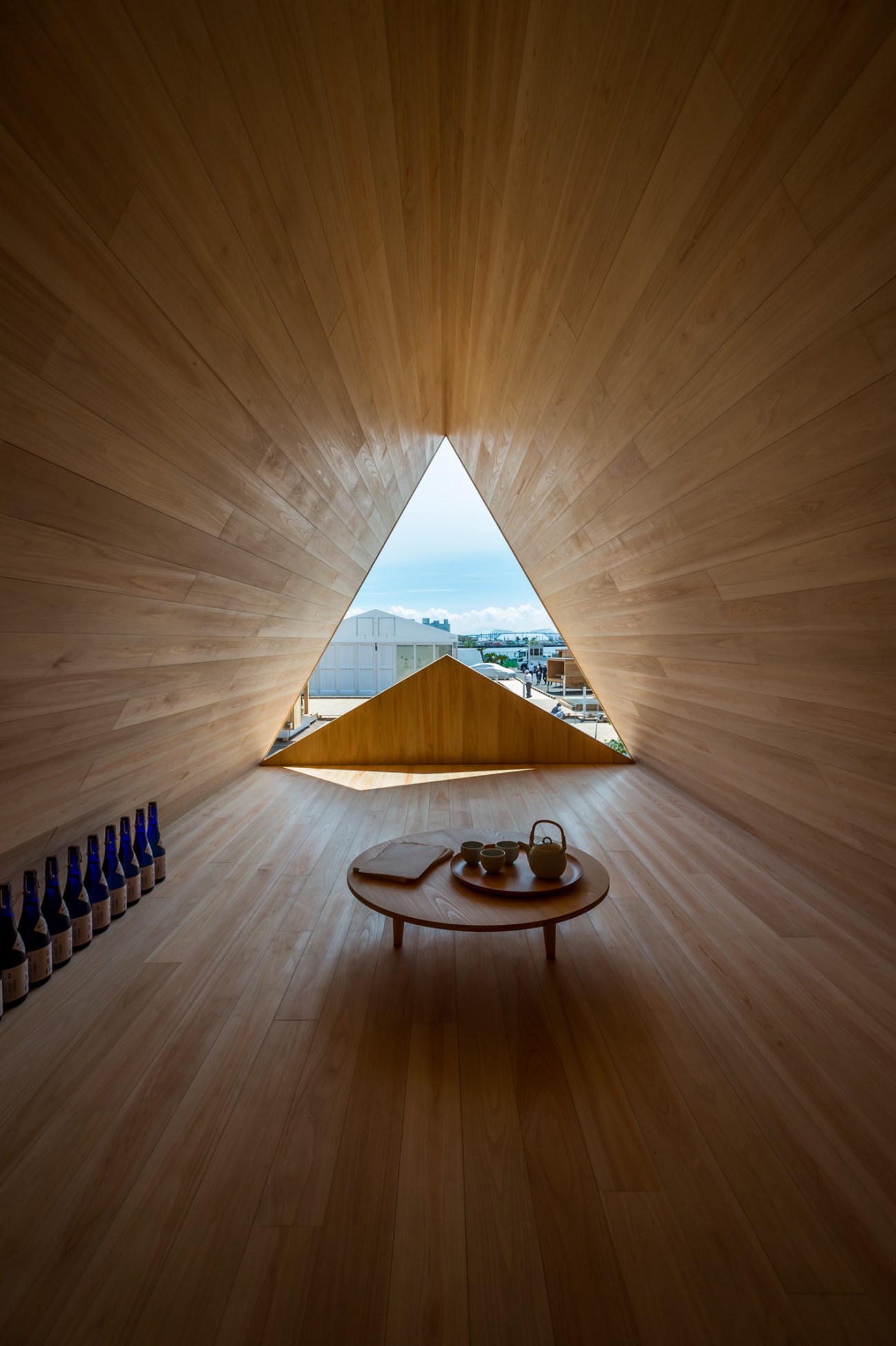 Interior of Yoshino-sugi Cedar House with triangular wooden frame window overlooking bay, round wooden table with tea service below.