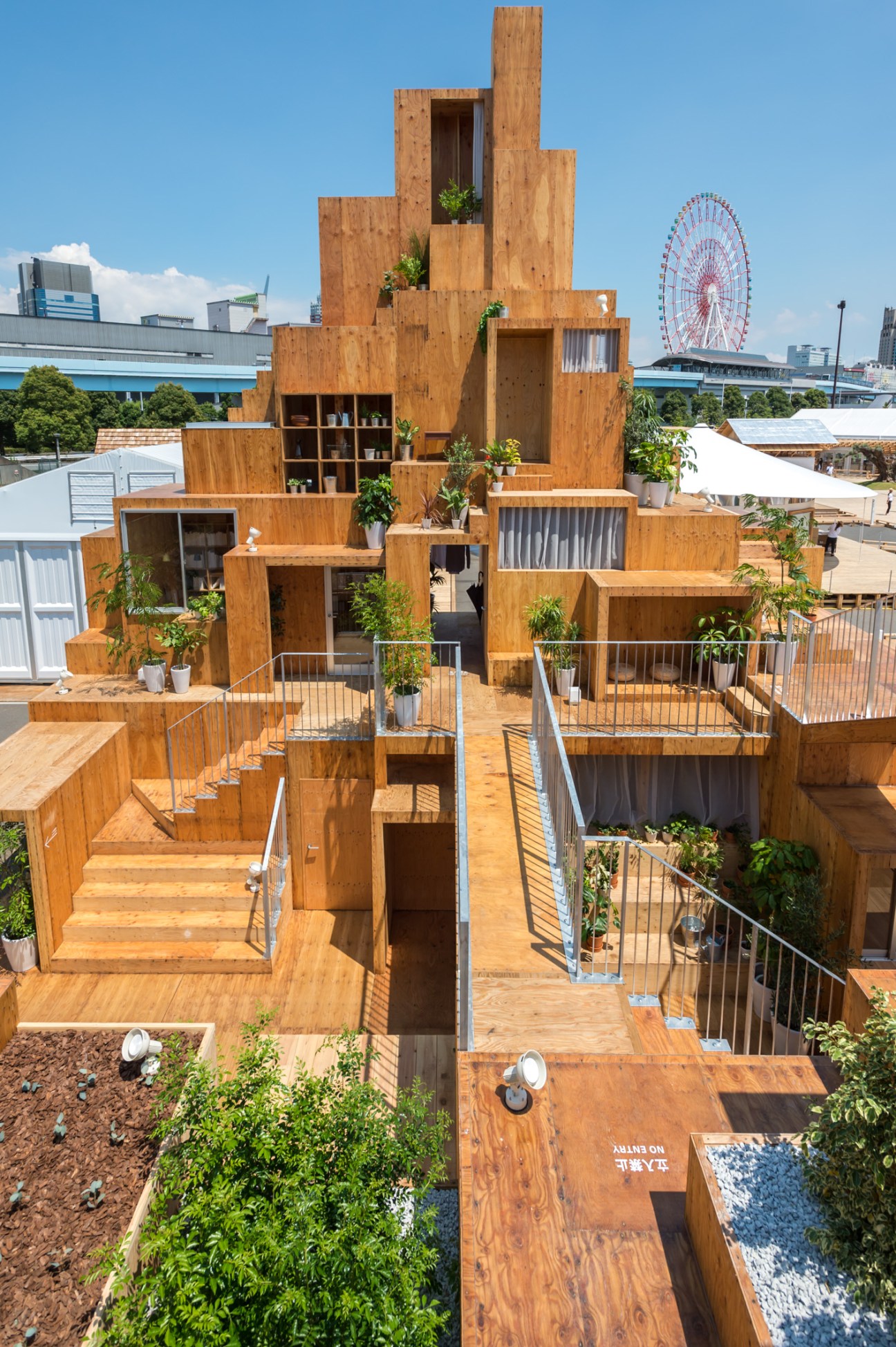 Wooden modular tower structure with stacked levels, open spaces, plants, and metal railings in an urban setting.