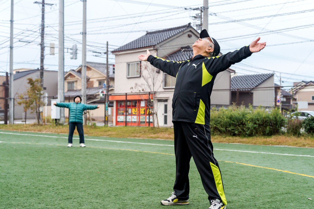 Elderly man stretching with arms outstretched during morning exercise at a sports field in Toyama, Japan.