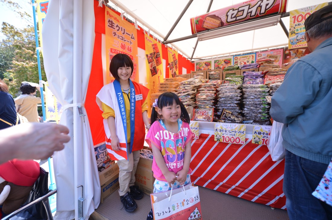 Young girl in pink shirt smiling at a confectionery expo stall stacked with packaged sweets and candies.