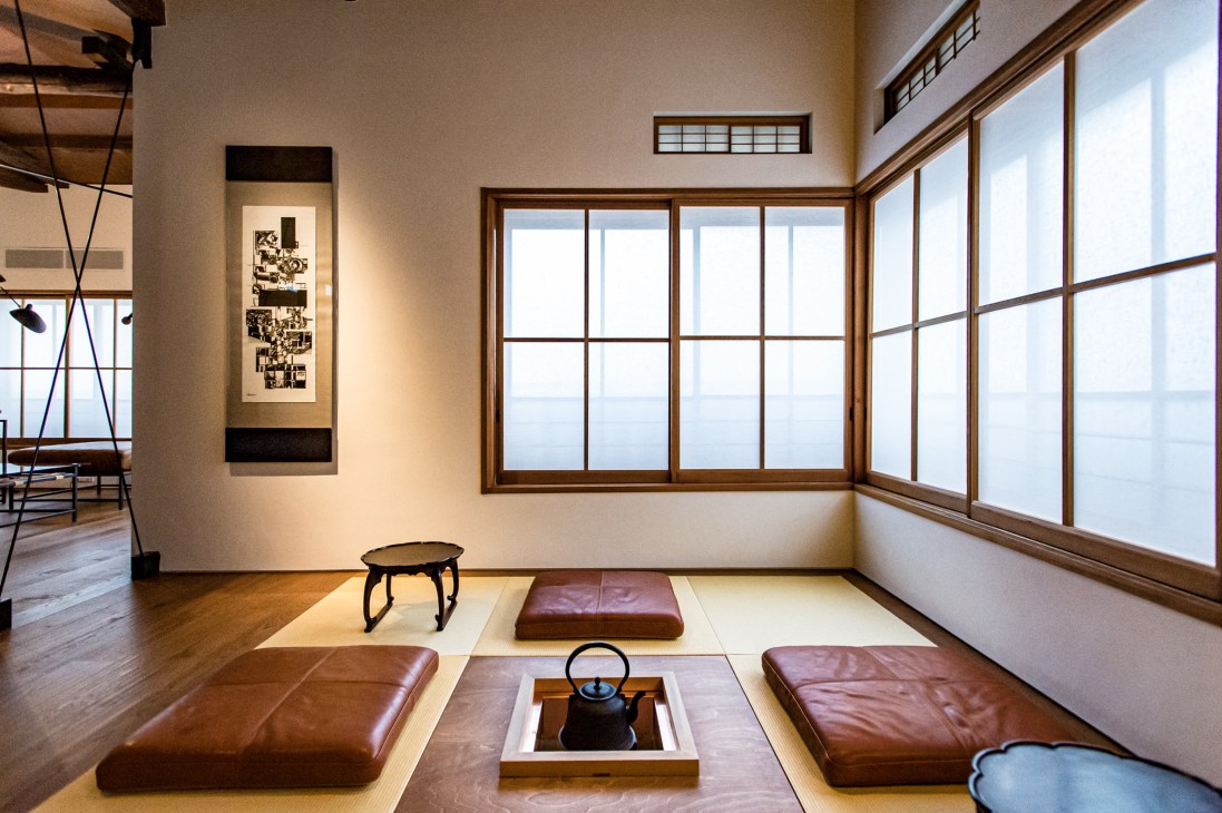Sunlit Japanese room with tatami mats, leather cushions, small tables, and large windows with wooden frames overlooking water.