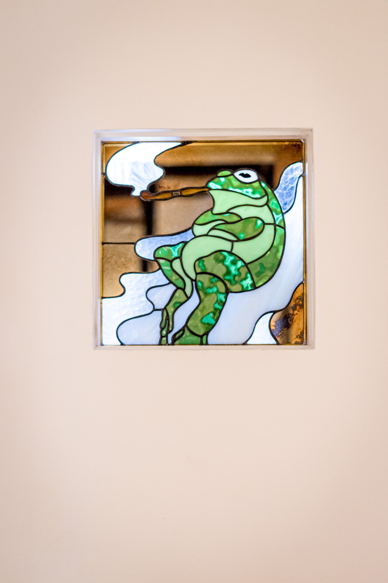 A stained-glass artwork depicting a green frog with blue and white accents in a brass-framed panel.