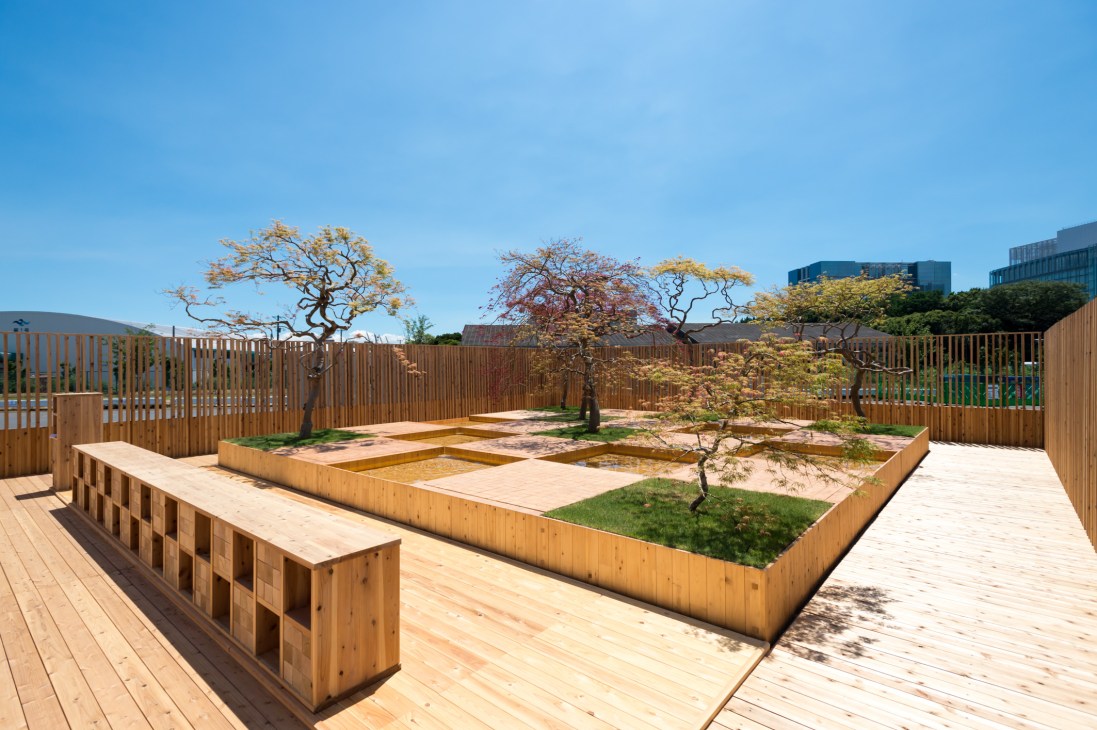 Wooden deck and garden with checkerboard layout featuring raised planting beds, mature maple trees, and water features under clear blue sky.