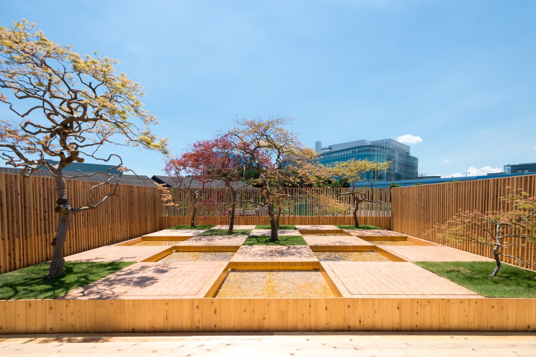 Checkerboard Water Garden with wooden platform paths, water channels, and Japanese maple trees with yellow and red foliage, enclosed by wooden fencing with modern buildings visible beyond.