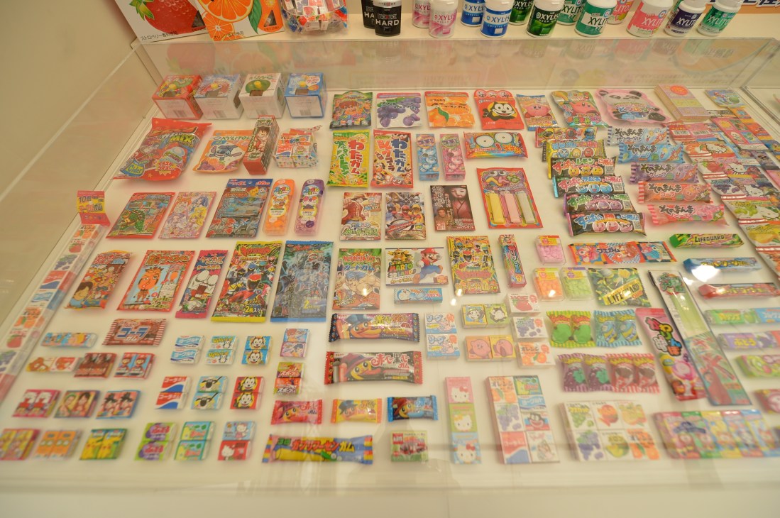 Display case of colorful Japanese chewing gum packets and candy boxes arranged on white shelving with bottles visible above.