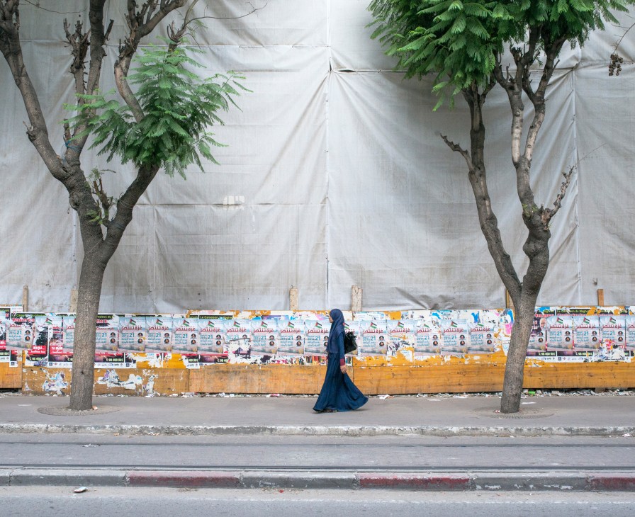 Woman in blue niqab walking past campaign posters on a Tunis street lined with trees.