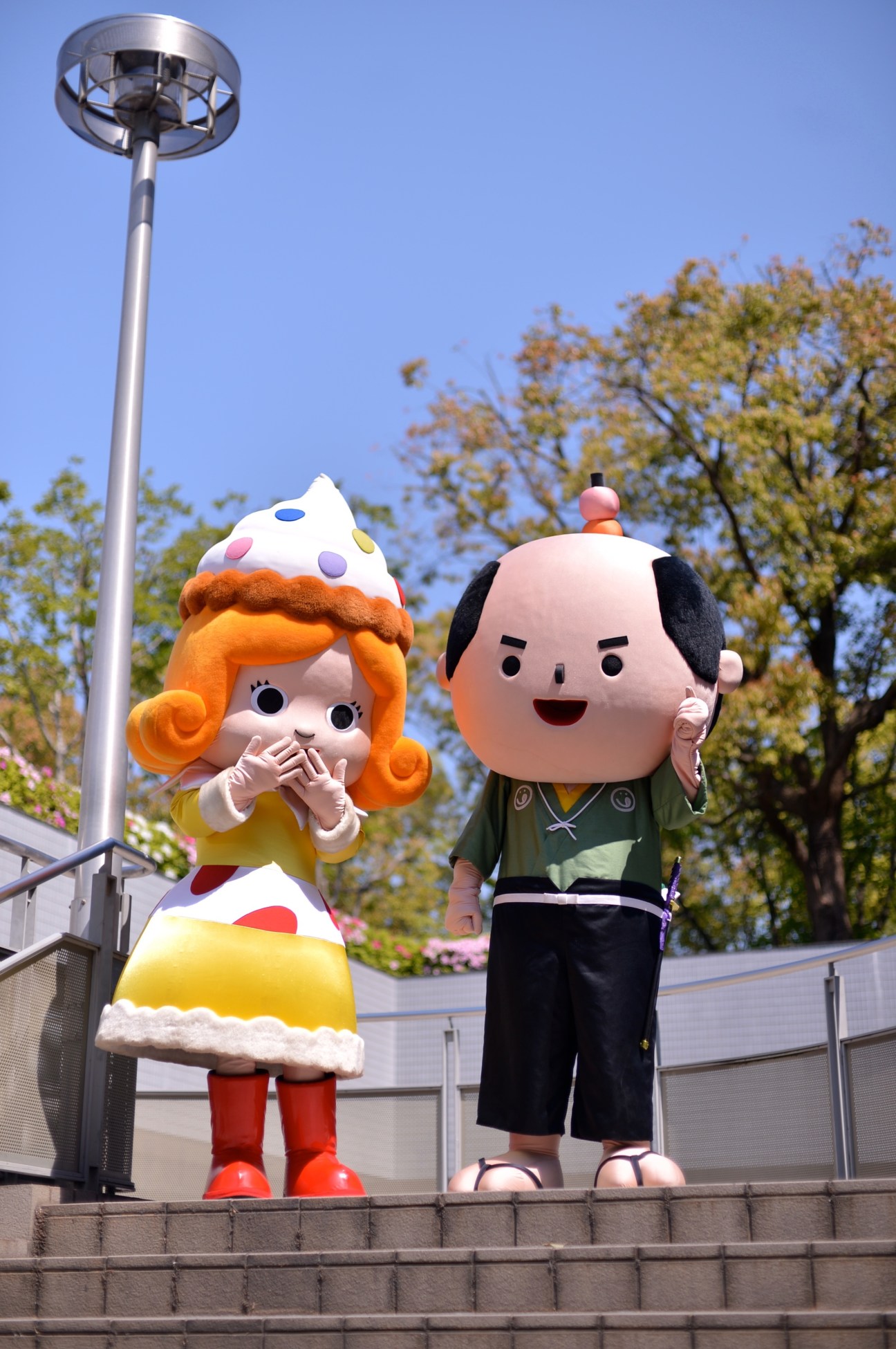 Two large colorful mascot characters: Kashinari-kun and Sweets-hime, standing outdoors at the confectionery expo.