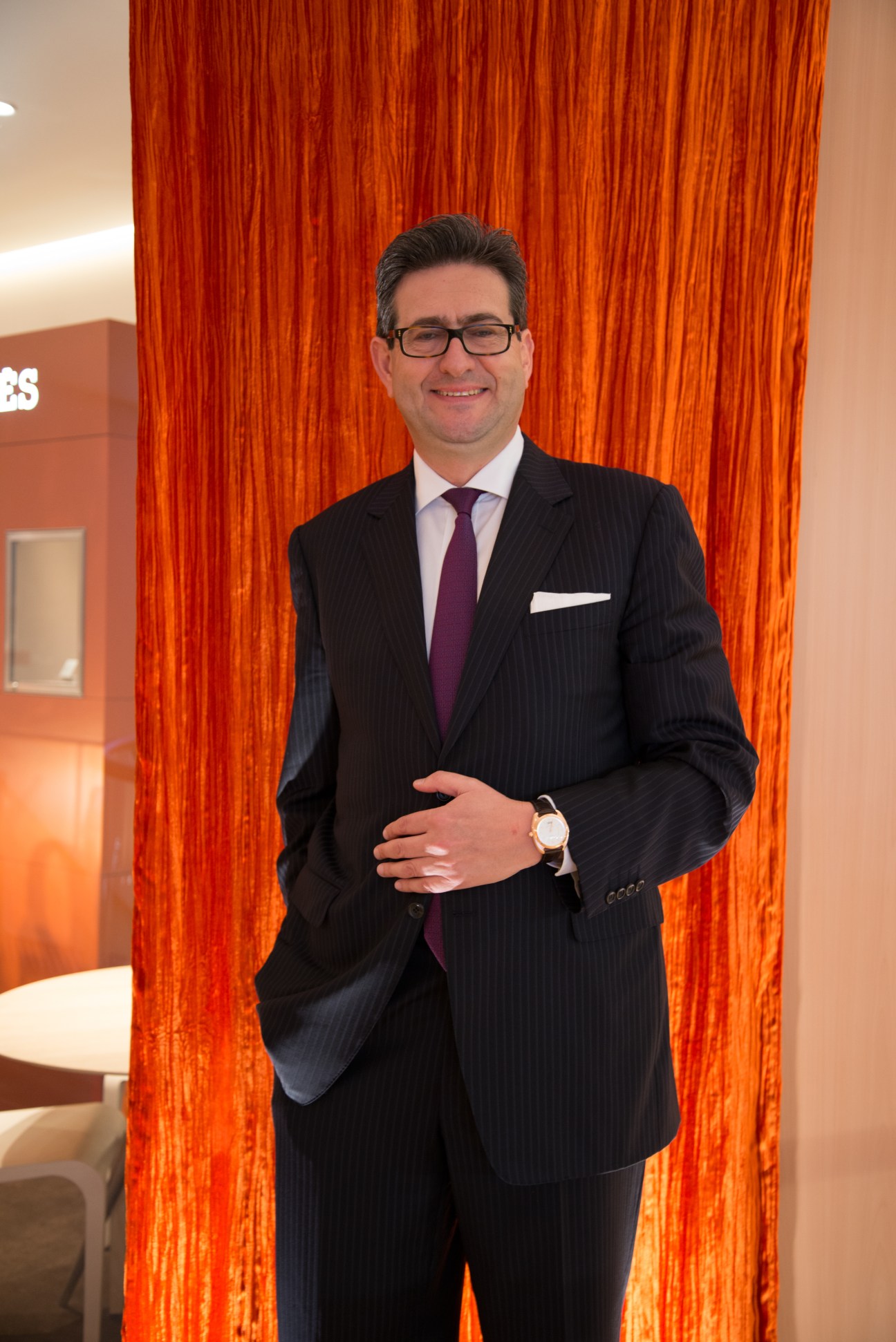 Luc Perramond, CEO of La Montre Hermès, stands in a dark pinstriped suit against a vibrant orange textured backdrop.